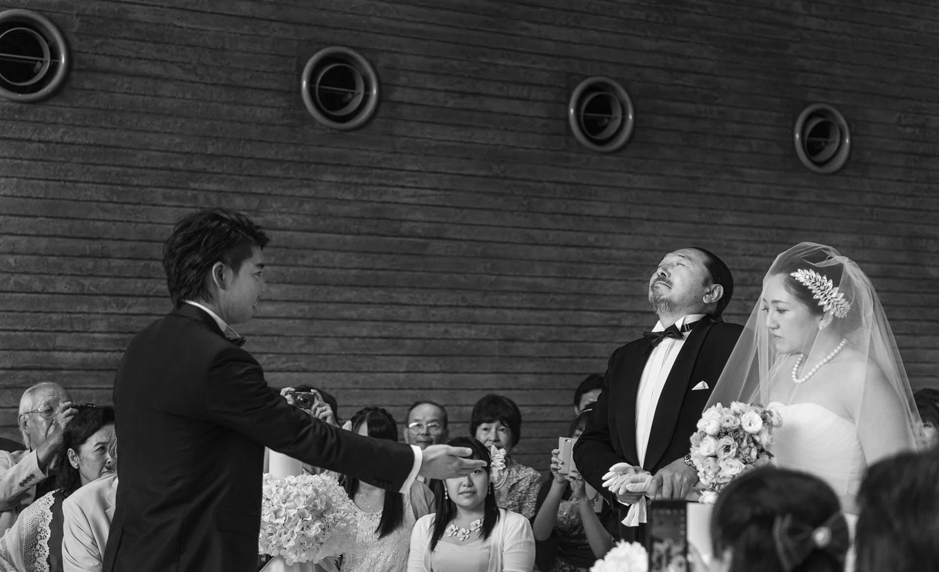 In black and white, the bride and groom stand at the front of their ceremony as guests look on in Yokohama.